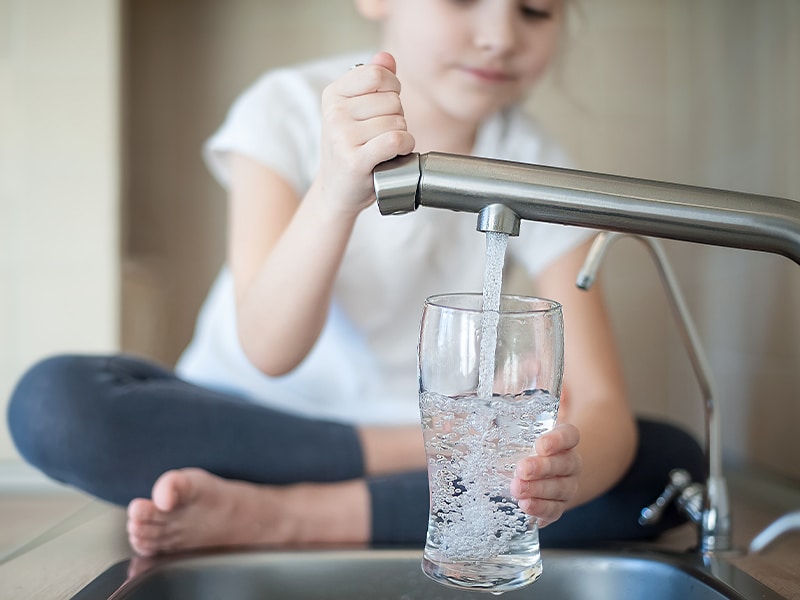 well-water-installations-and-service Child fills glass from kitchen faucet while sitting on counter, emphasizing the value of well water testing for safe drinking.